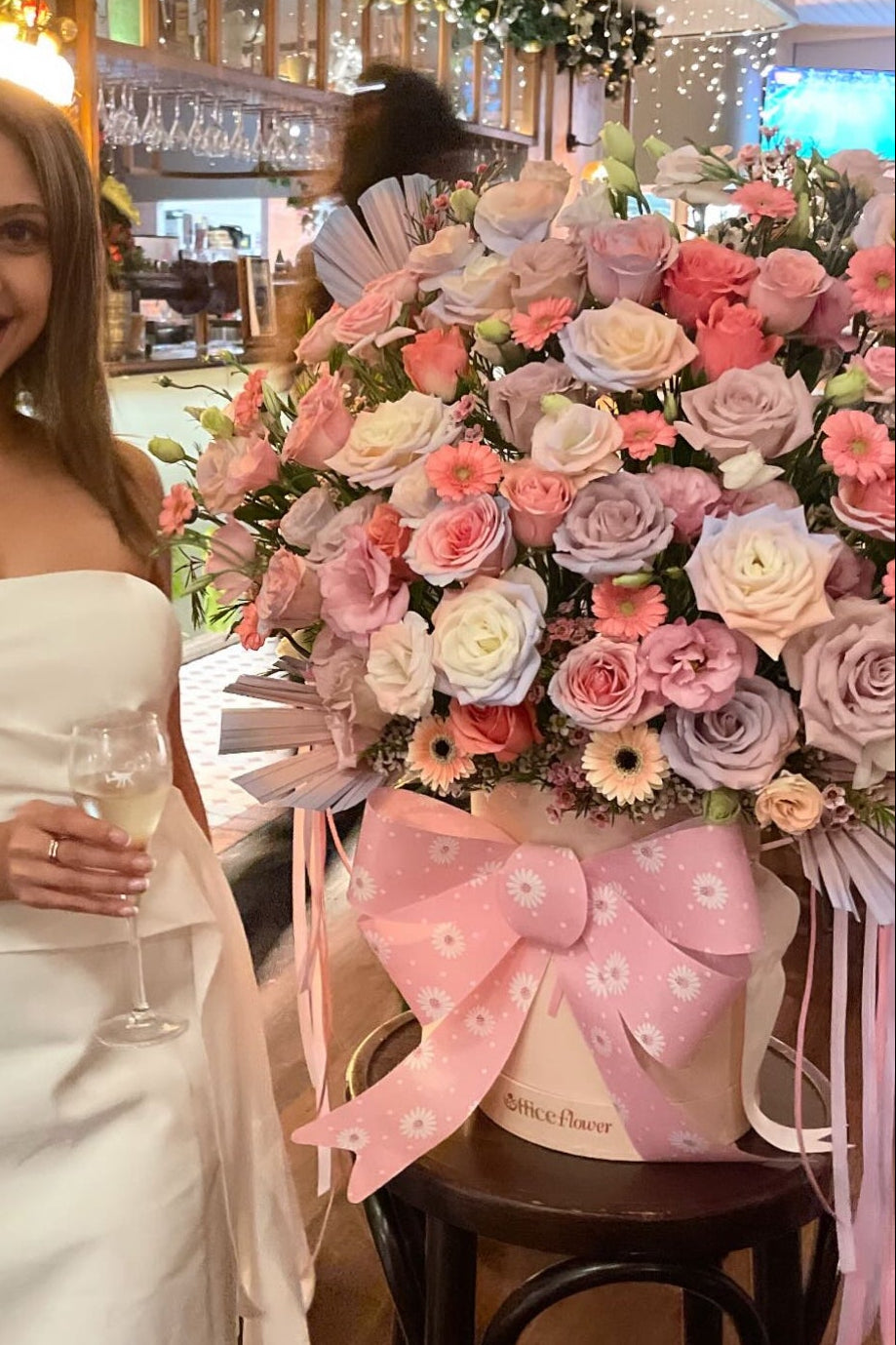 Woman in a white dress standing next to a large floral arrangement with a pink bow indoors.