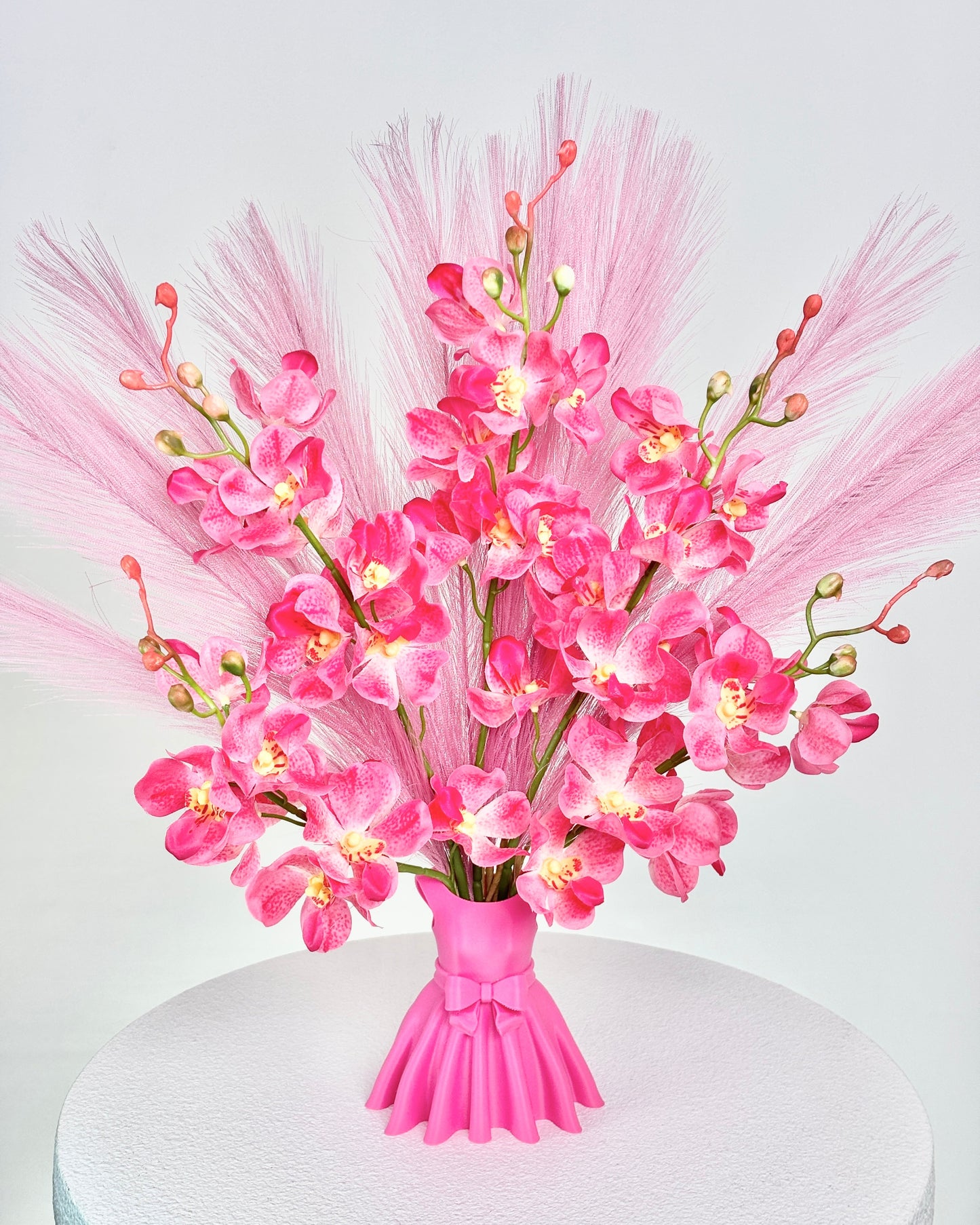 Artificial flower arrangement featuring bright pink orchids and soft pink pampas grass, beautifully styled in a sculptural pink vase shaped like an elegant dress, placed on a round white table against pink room background.