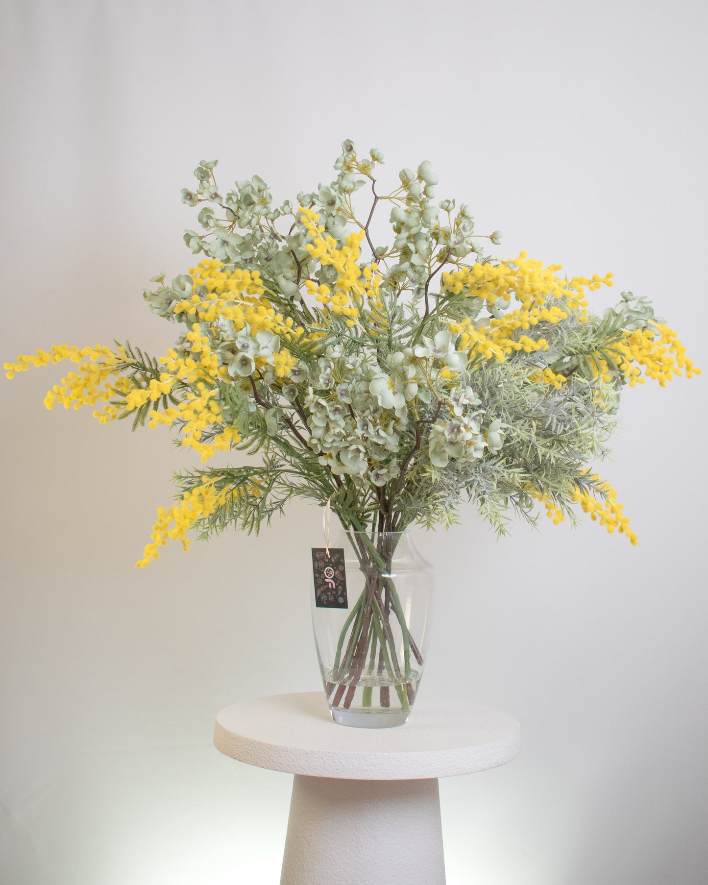 Artificial flower arrangement featuring vibrant yellow wattle blooms mixed with soft green eucalyptus leaves, displayed in a clear glass vase filled with faux stems for a natural, elegant look.