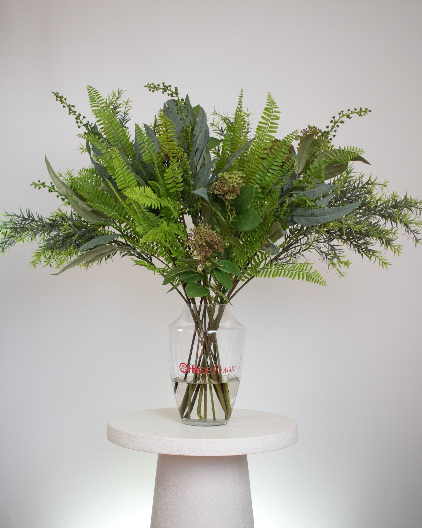 Artificial green foliage arrangement with ferns and leafy stems, displayed in a clear glass vase with acrylic water