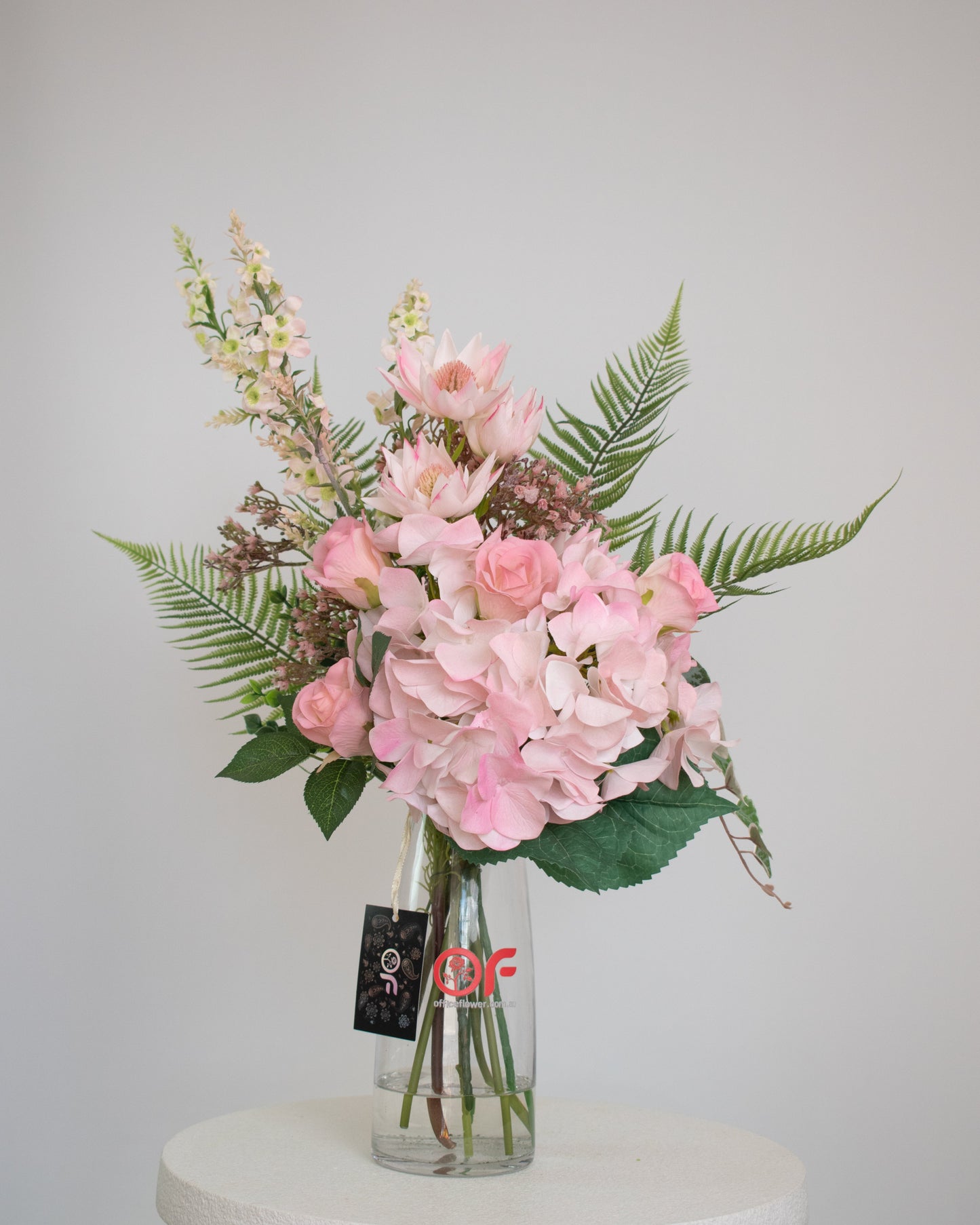 Artificial pink floral arrangement with hydrangeas, roses, and ferns, arranged in a clear glass vase with acrylic water.