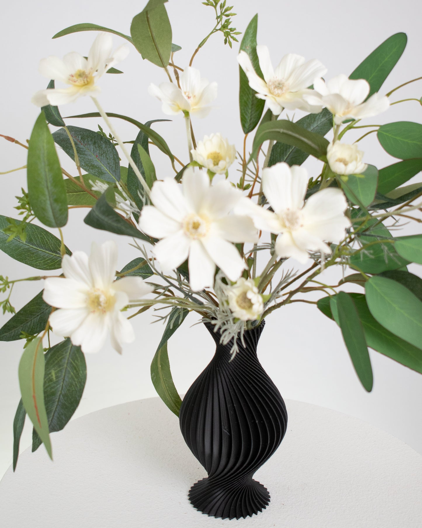 Artificial arrangement featuring white daisy-like flowers and lush eucalyptus leaves in a sculpted matte black vase, styled on a white pedestal — perfect for modern, minimalist, or monochrome interiors.