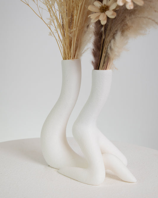 A modern dried floral arrangement featuring pampas grass, bunny tails, preserved foliage, and neutral-toned dried flowers, artistically displayed in a sculptural white ceramic double vase shaped like two flowing figures.