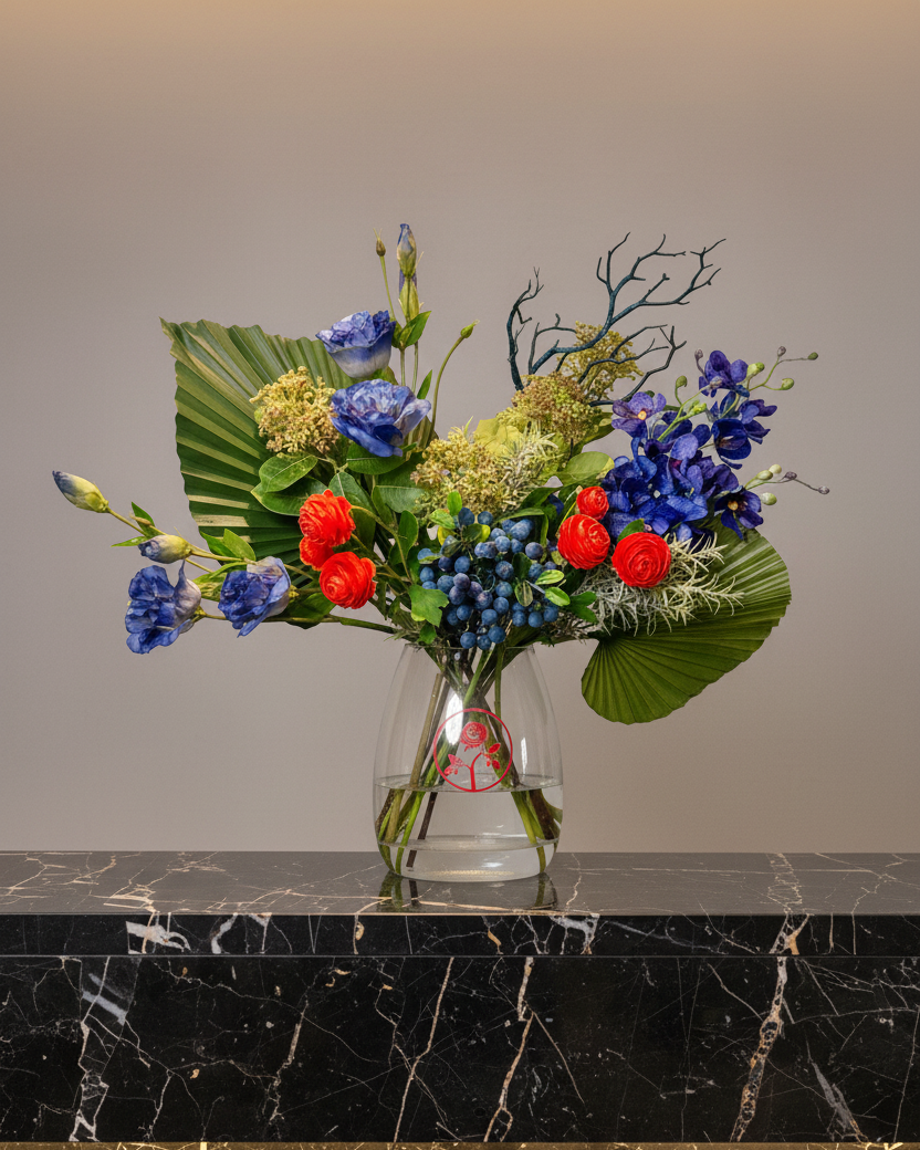 A vibrant artificial floral arrangement in a clear glass vase, set on a black marble surface against a warm beige background.