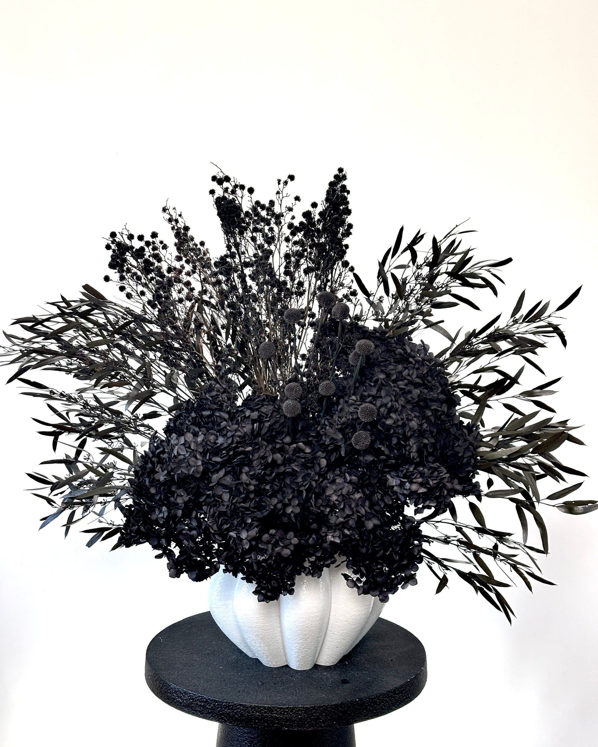 A black dried floral arrangement featuring dense black baby's breath and silver-gray olive leaves, presented in a textured white ribbed vase on a round black pedestal against a plain white background.