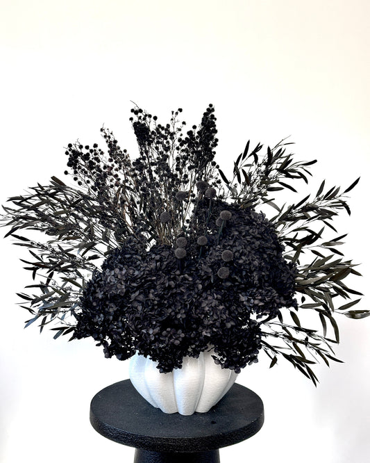 A black dried floral arrangement featuring dense black baby's breath and silver-gray olive leaves, presented in a textured white ribbed vase on a round black pedestal against a plain white background.