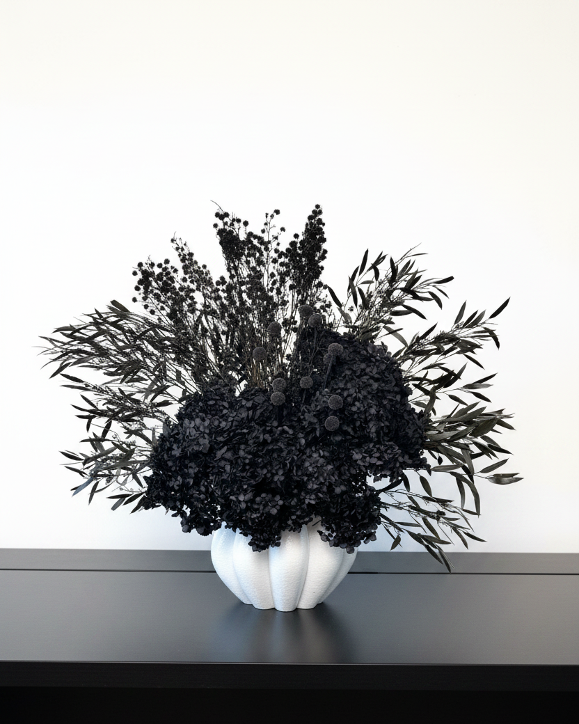 A black dried floral arrangement in a white ribbed ceramic vase, displayed on a dark shelf against a plain white wall.