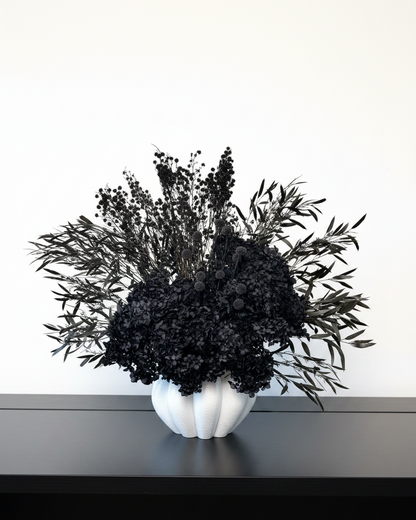 A black dried floral arrangement in a white ribbed ceramic vase, displayed on a dark shelf against a plain white wall.