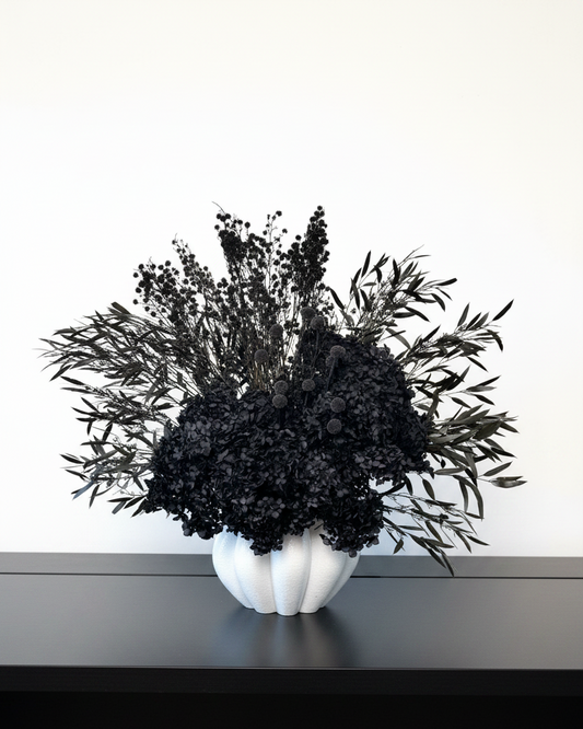 A black dried floral arrangement in a white ribbed ceramic vase, displayed on a dark shelf against a plain white wall.