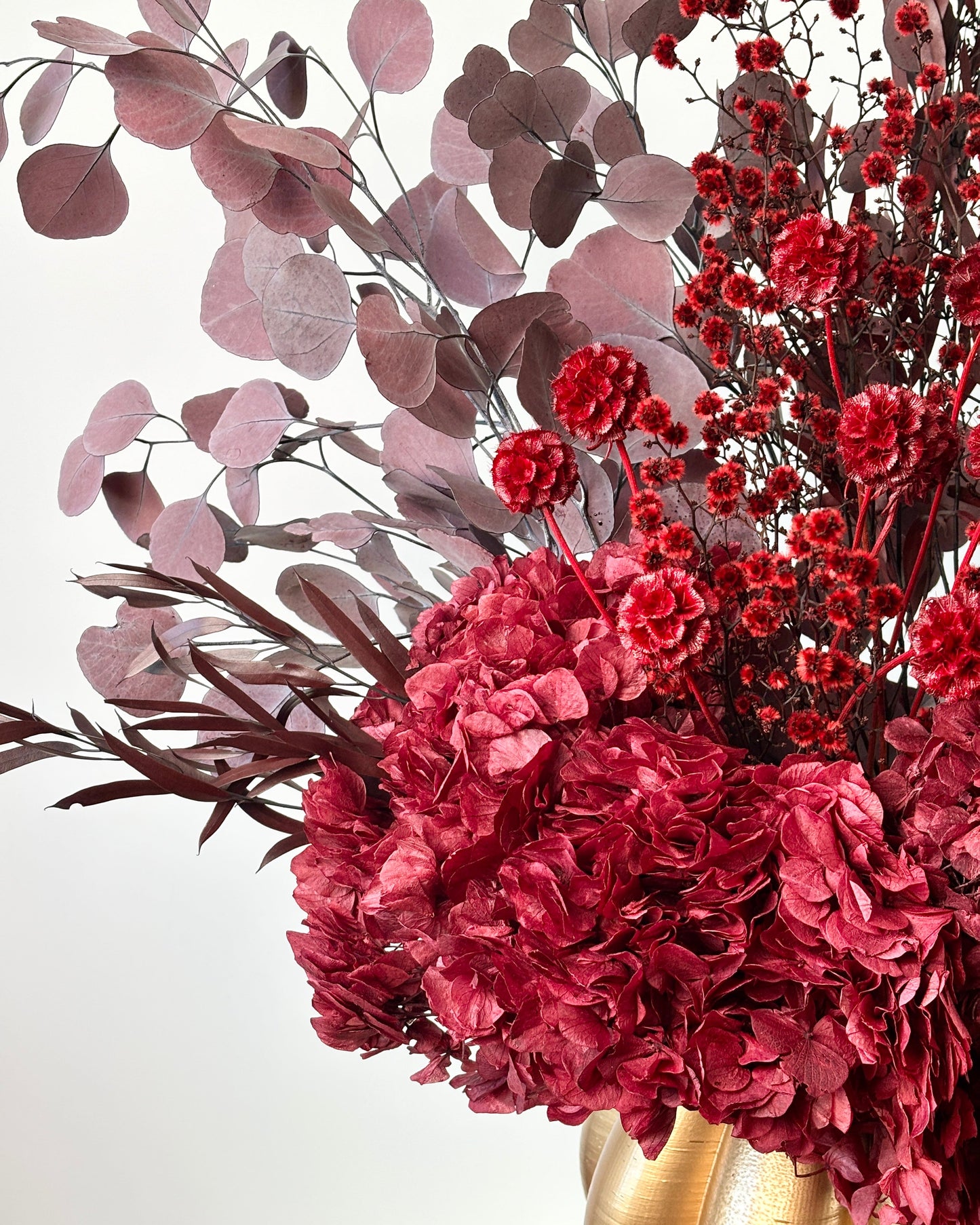 A decorative floral arrangement featuring deep pink hydrangeas, red berries, dusty rose eucalyptus, and dark burgundy stems in a shiny gold vase against a clean white background.
