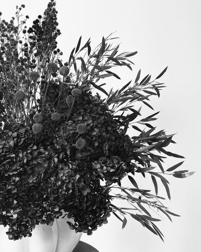 A black and white photograph of a dried floral arrangement with textured spherical blooms and long narrow leaves, set against a plain light background.