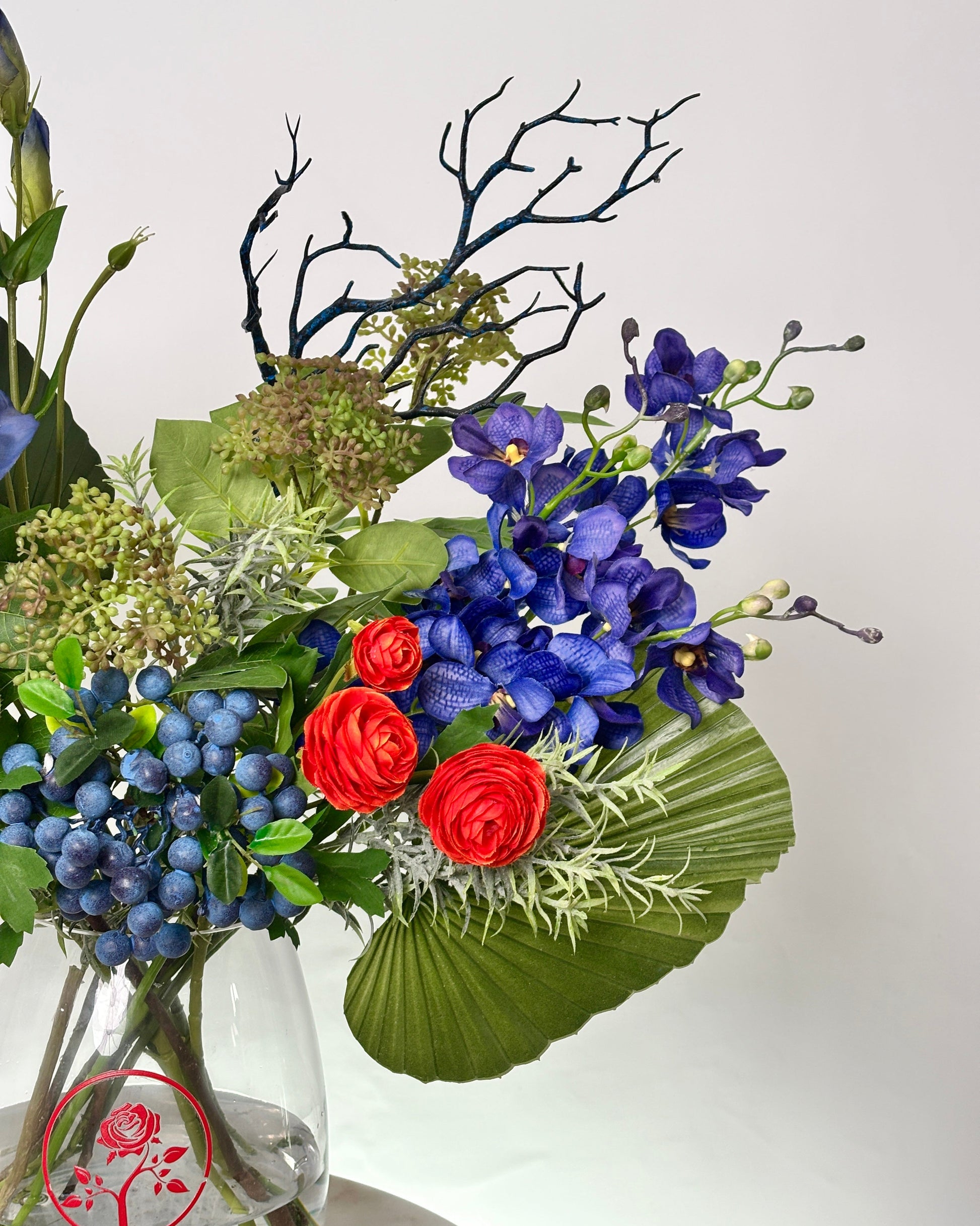 A floral arrangement in a clear glass vase featuring vivid blue orchids, bright red roses, blue berries, green foliage, and dark branch elements against a plain light background.