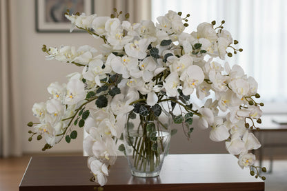 White orchid arrangement in a clear vase on a wooden table with a blurred indoor background
