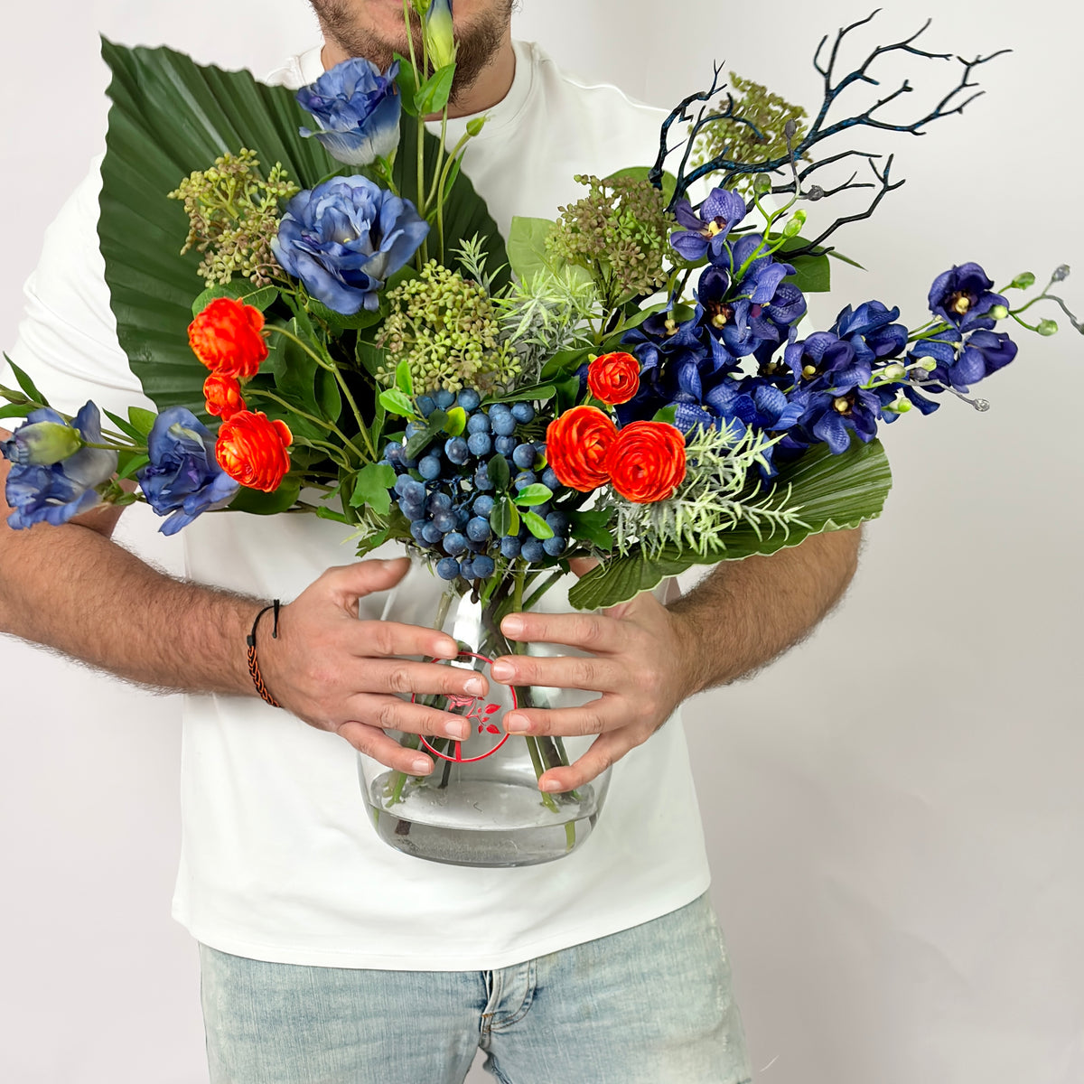 A person in a white t-shirt and light blue jeans holds a large, vibrant mixed floral arrangement in a clear glass vase against a plain white background.