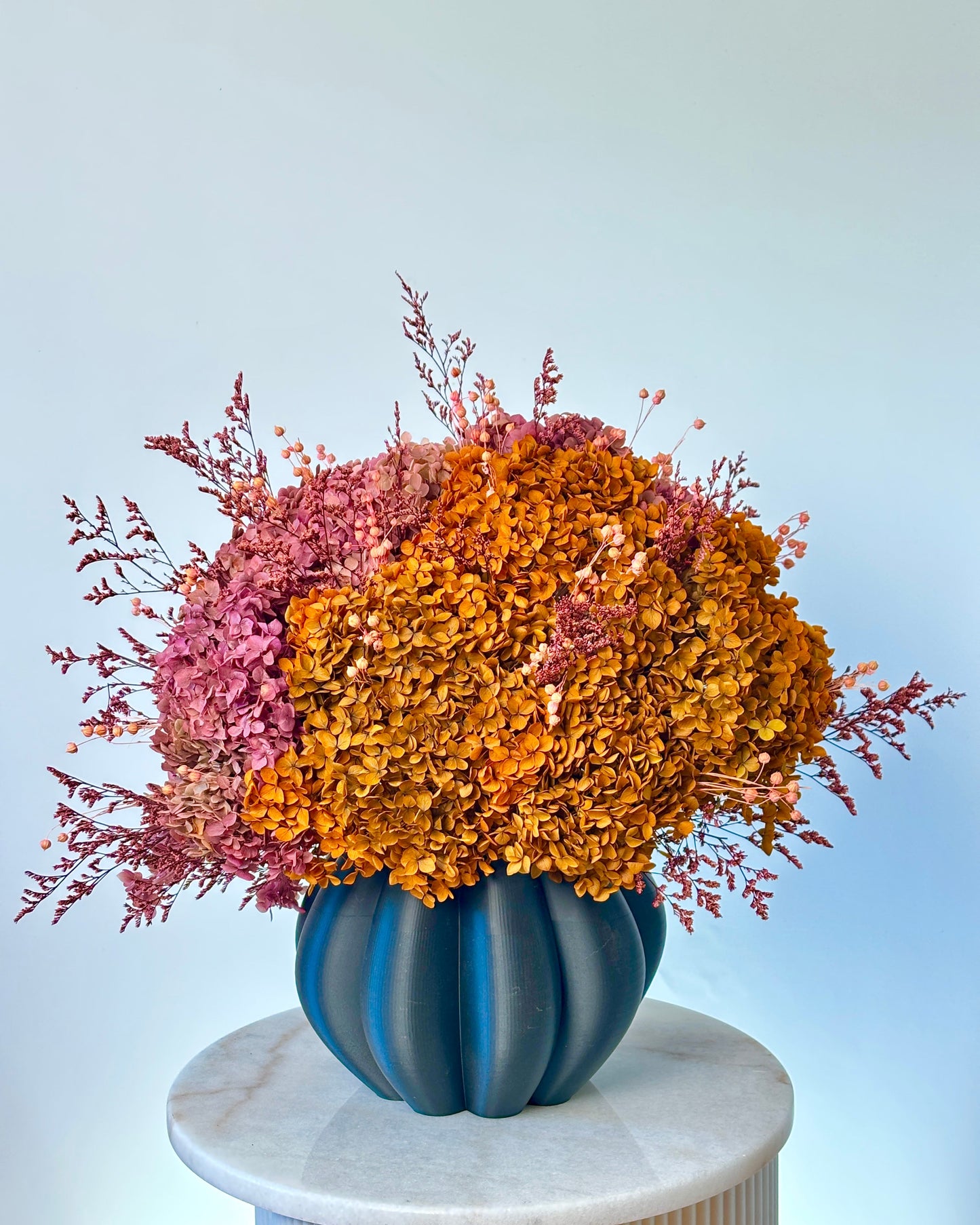 A dried floral arrangement with dense clusters of orange, mustard, and dusty pink botanicals in a ribbed dark blue vase, displayed on a white marble pedestal against a pale blue background.
