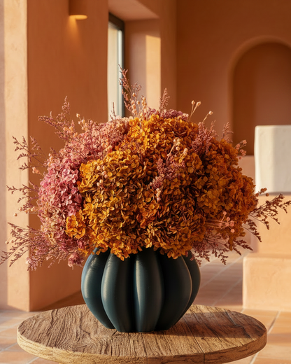 A dark teal ribbed ceramic vase filled with a large dried floral arrangement in orange, rust, and dusty pink tones, displayed on a round wooden table in a warm terracotta-toned interior room.
