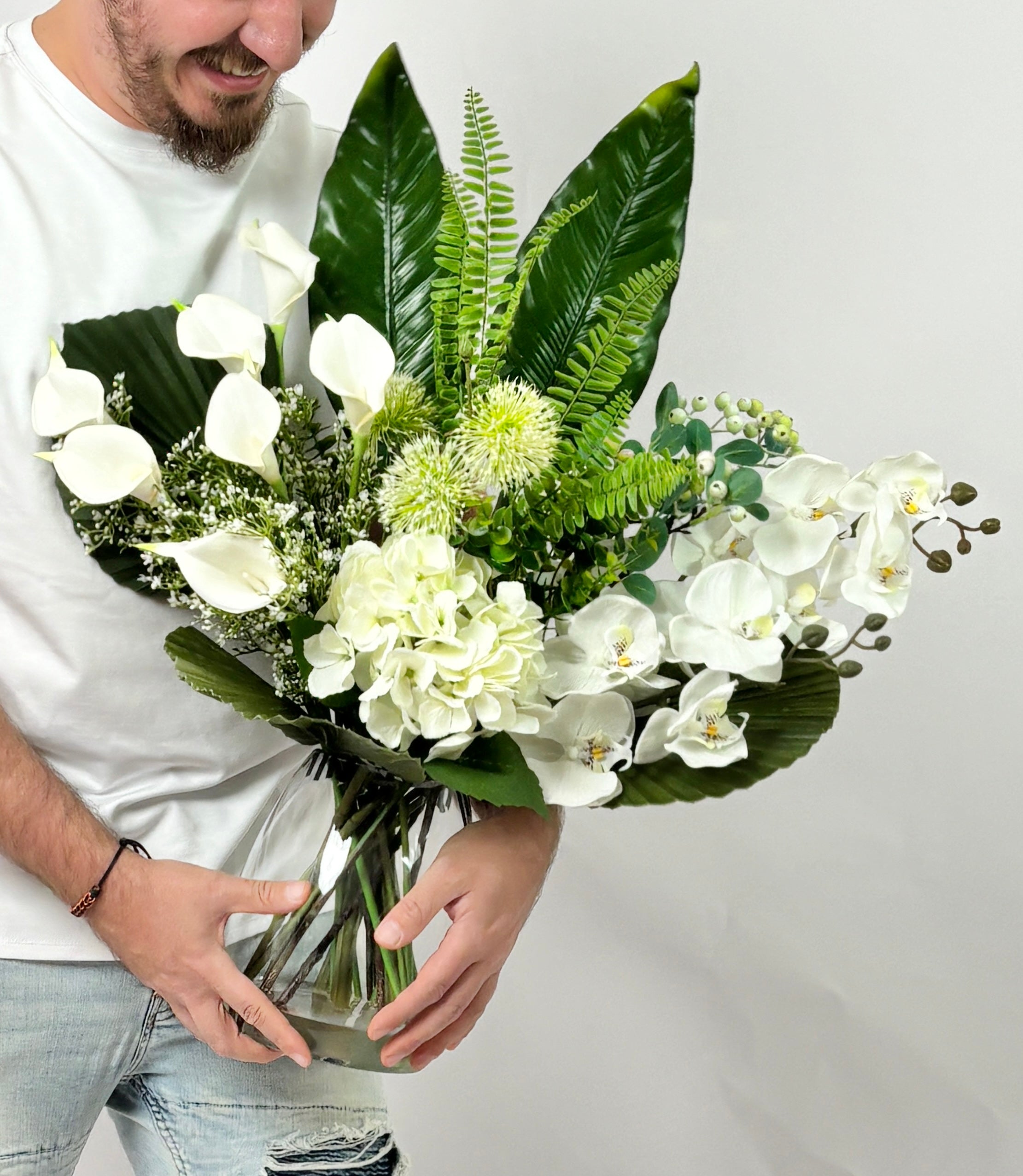 A man in a white t-shirt and jeans holds a large, vibrant artificial floral arrangement in a clear glass vase. The bouquet features white calla lilies, white orchids, white hydrangeas, green ferns, and large green tropical leaves against a plain light gray background.