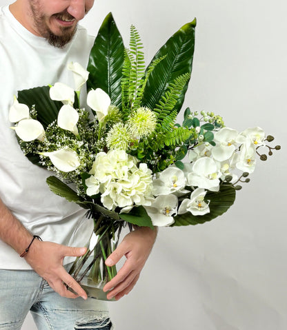 A man in a white t-shirt and jeans holds a large, vibrant artificial floral arrangement in a clear glass vase. The bouquet features white calla lilies, white orchids, white hydrangeas, green ferns, and large green tropical leaves against a plain light gray background.