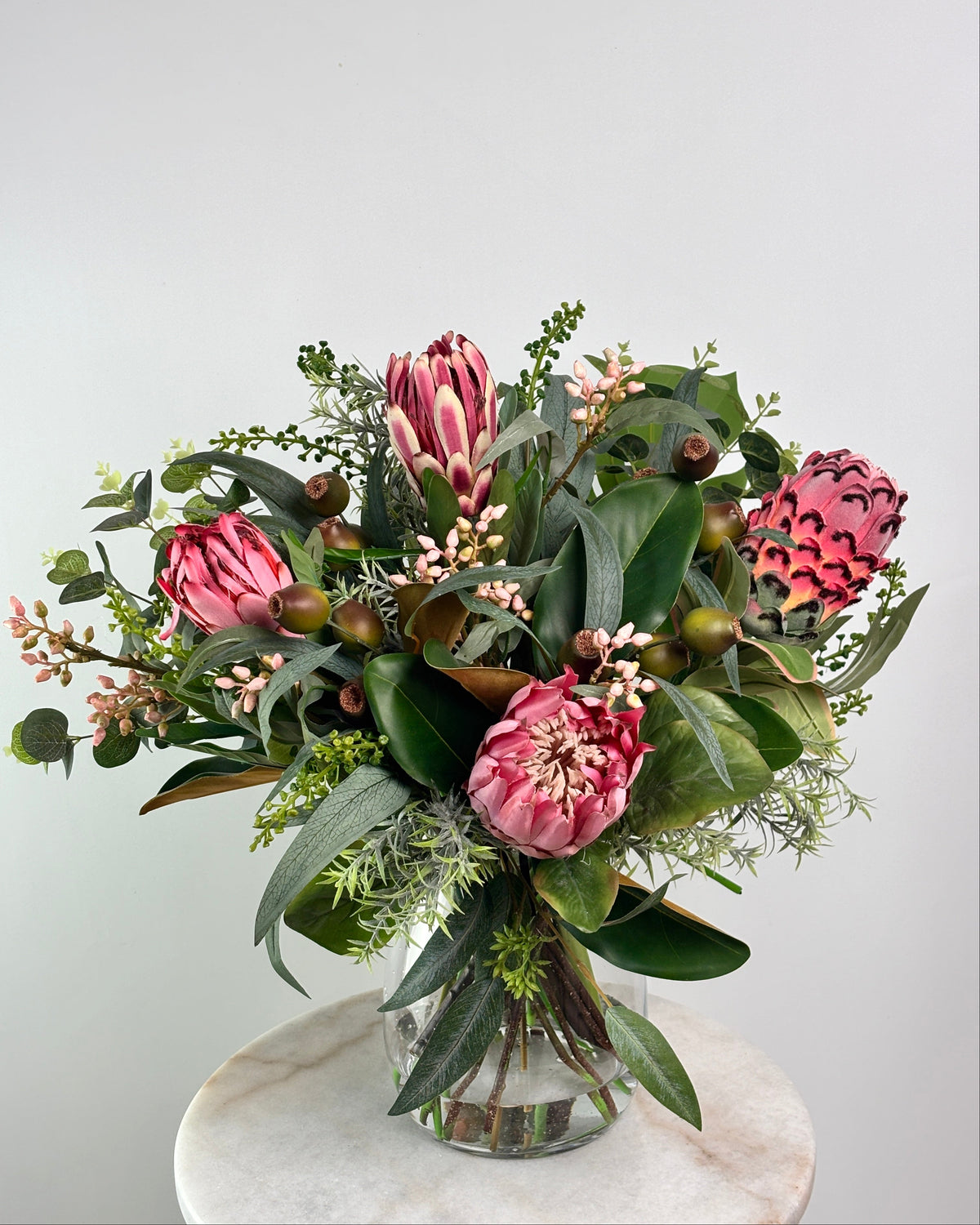 A vibrant artificial flower bouquet featuring prominent pink protea-like blooms, glossy green leaves, and small pale pink filler flowers, arranged in a clear glass vase on a round white marble surface against a light gray background.
