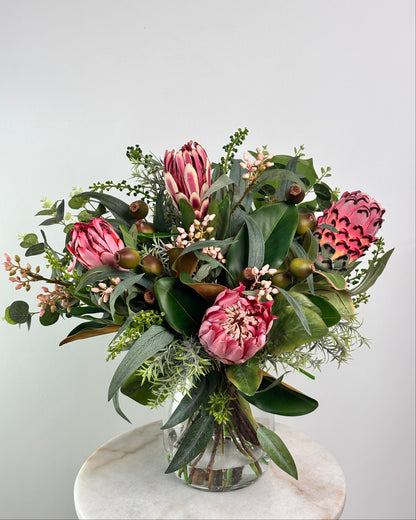 A vibrant artificial flower bouquet featuring prominent pink protea-like blooms, glossy green leaves, and small pale pink filler flowers, arranged in a clear glass vase on a round white marble surface against a light gray background.