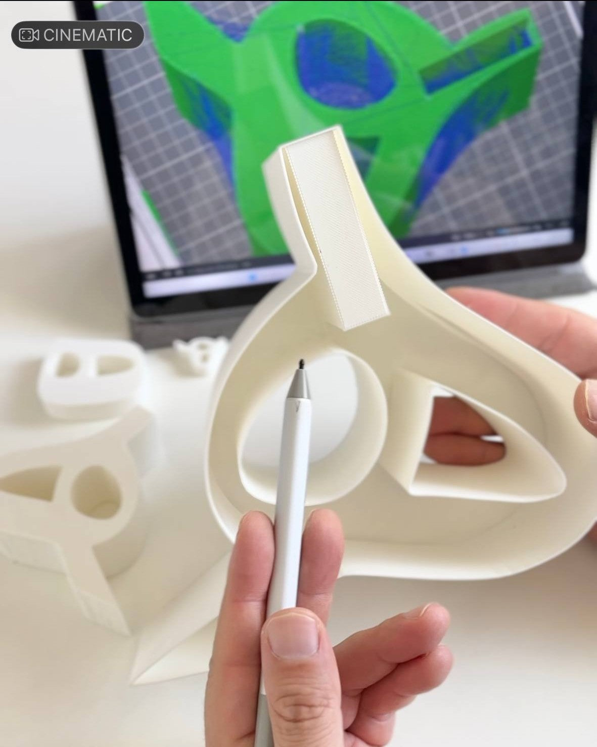 Hand holding a 3D-printed object with a laptop displaying 3D design in the background