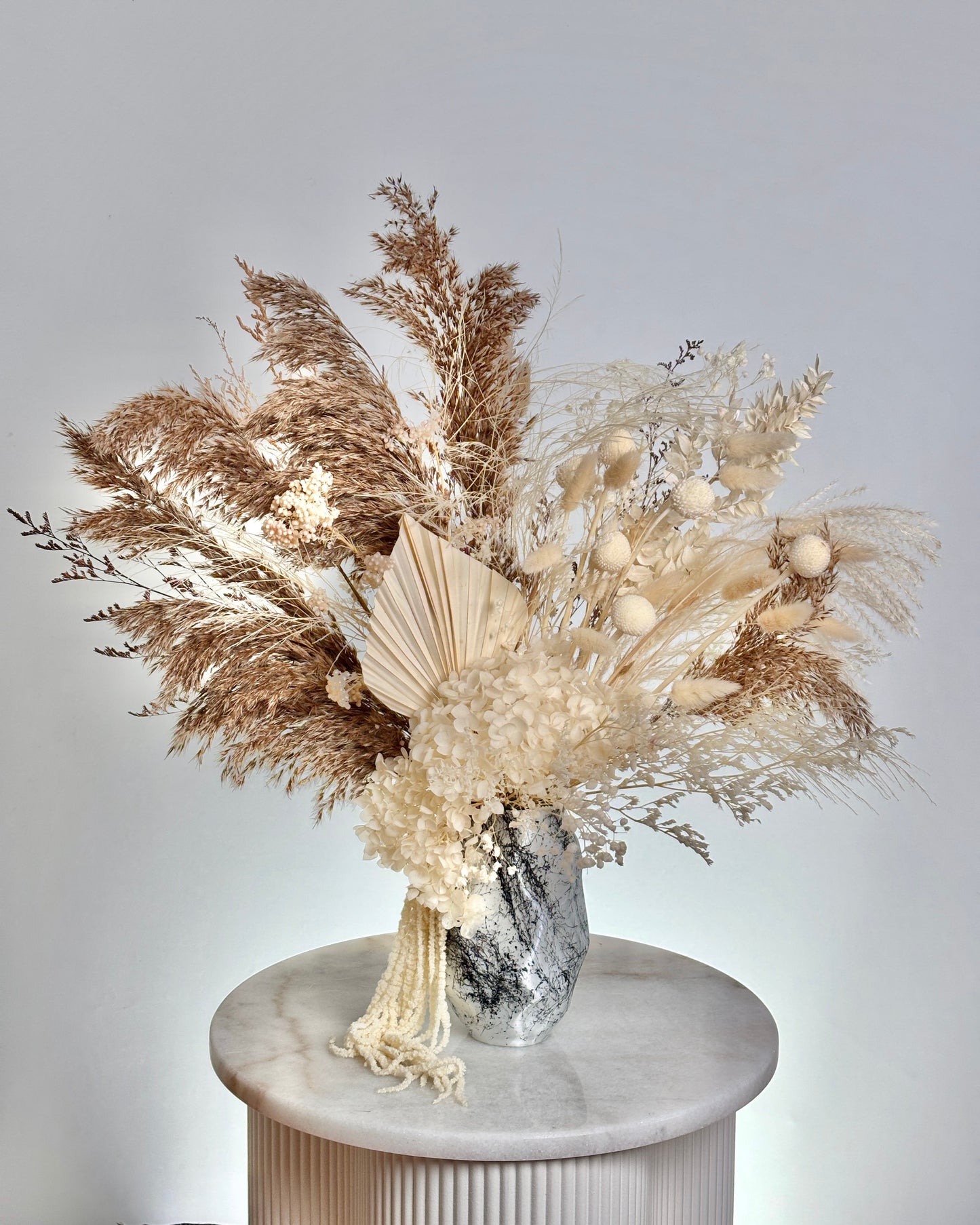 Decorative bouquet with dried pampas grass and white flowers in a silver vase on a white pedestal against a light gray background.