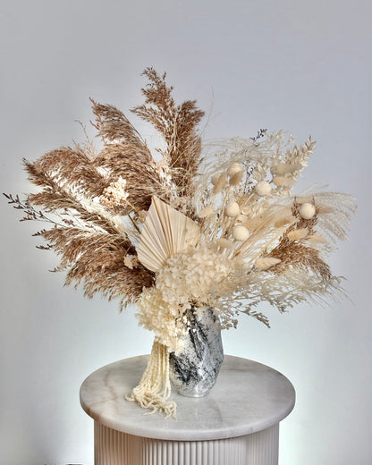 Decorative bouquet with dried pampas grass and white flowers in a silver vase on a white pedestal against a light gray background.