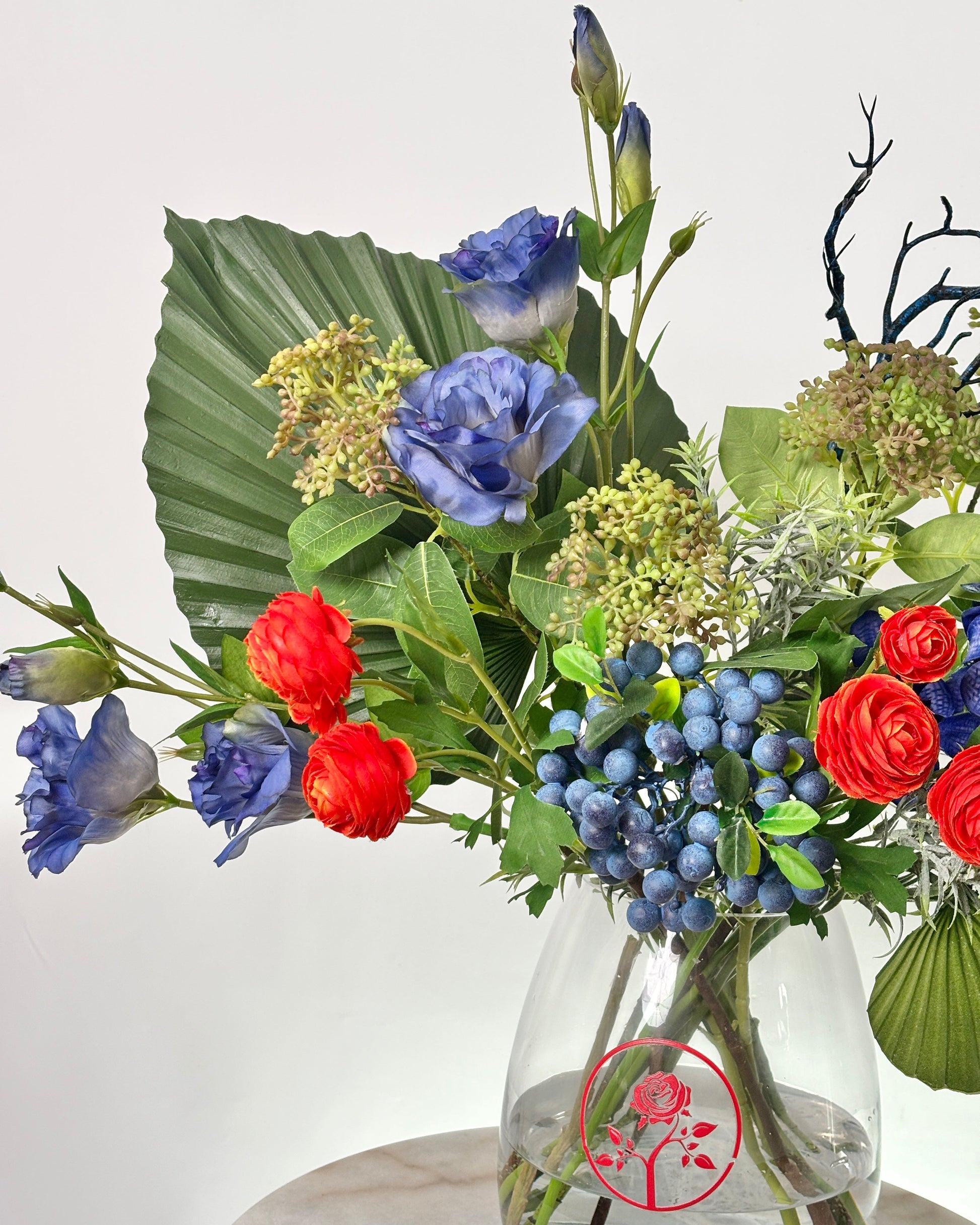 A vibrant artificial floral arrangement in a clear glass vase, featuring blue lisianthus, red roses, blue berries, and green foliage against a plain light background.