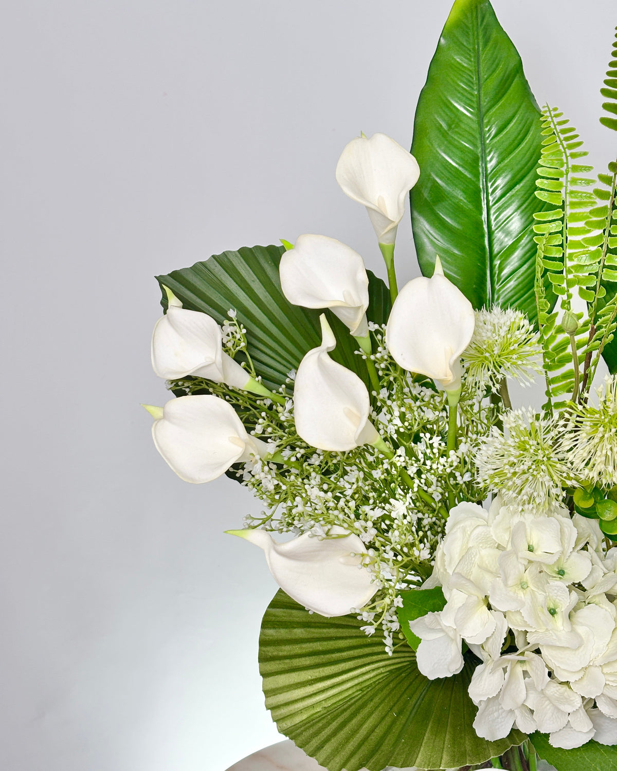A close-up of a floral arrangement featuring white calla lilies, white hydrangeas, green palm-leaf foliage, and green ferns against a light gray background