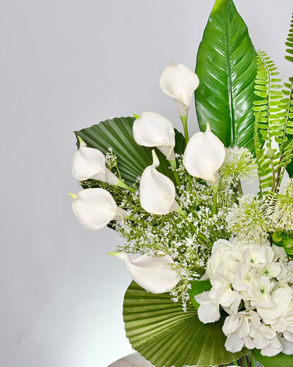 A close-up of a floral arrangement featuring white calla lilies, white hydrangeas, green palm-leaf foliage, and green ferns against a light gray background