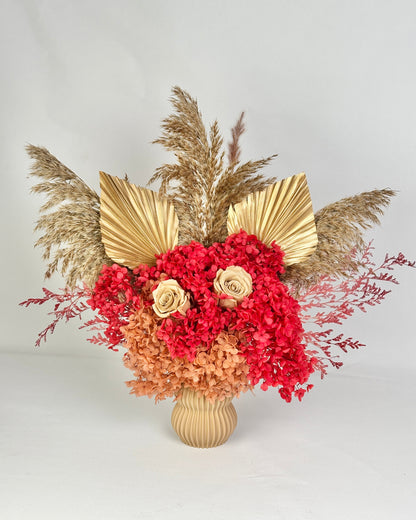 Decorative dried floral arrangement with red and gold flowers, pampas grass, and a textured vase on a light gray background.