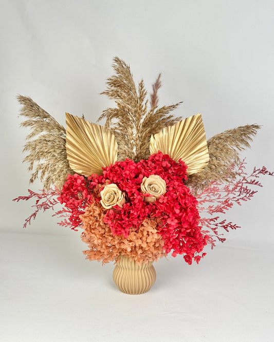 Decorative dried floral arrangement with red and gold flowers, pampas grass, and a textured vase on a light gray background.