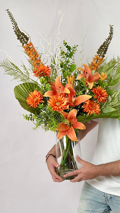 A vibrant orange and green floral arrangement in a clear glass vase, held by a person wearing a white t-shirt and light blue jeans against a plain light gray background.