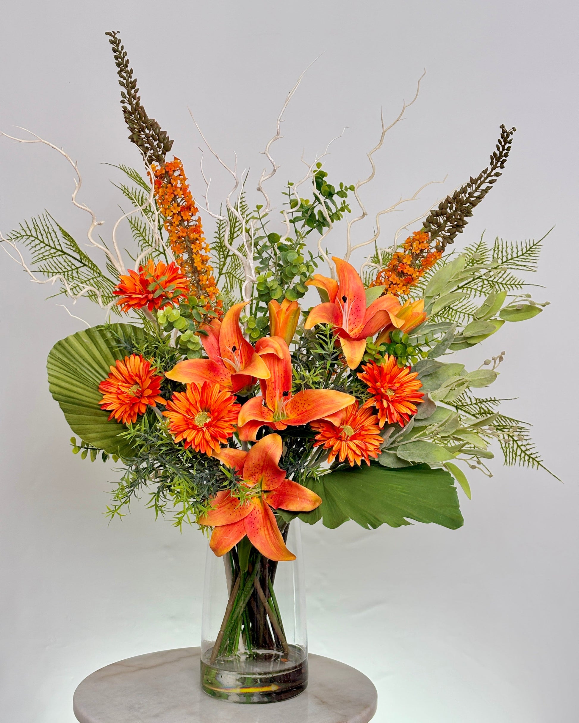 An front desk artificial floral arrangement in a clear glass vase, featuring bright orange lilies, orange daisies, green foliage, and brown ornamental grasses, displayed on a light gray marble-topped surface against a light gray background