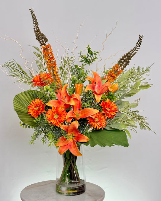 An front desk artificial floral arrangement in a clear glass vase, featuring bright orange lilies, orange daisies, green foliage, and brown ornamental grasses, displayed on a light gray marble-topped surface against a light gray background