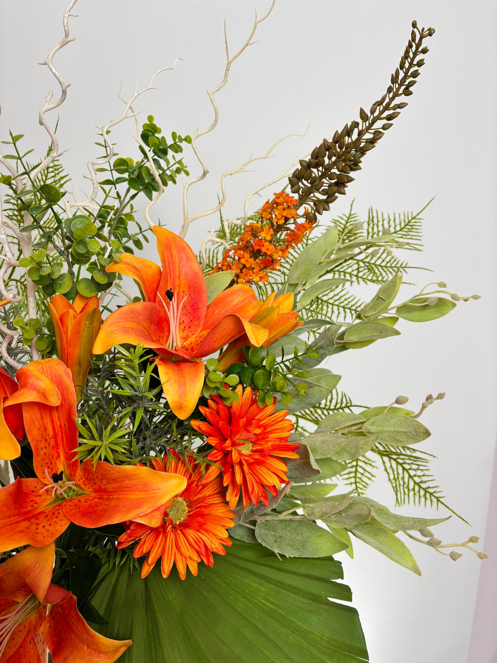 A close-up of a floral arrangement featuring bright orange lilies, orange gerbera daisies, green foliage, and dried botanicals against a plain light background.