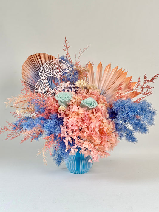 Preserved flower arrangement in pink, blue, and peach tones in a turquoise vase