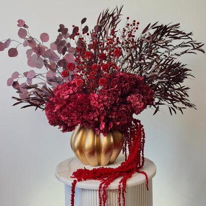A decorative floral arrangement in a ribbed metallic gold vase, displayed on a white pedestal against a plain light background. The arrangement features deep red hydrangeas, burgundy berries, dark burgundy feathery stems, and dusty pink round leaves, with red beaded garlands draped over the vase and pedestal.