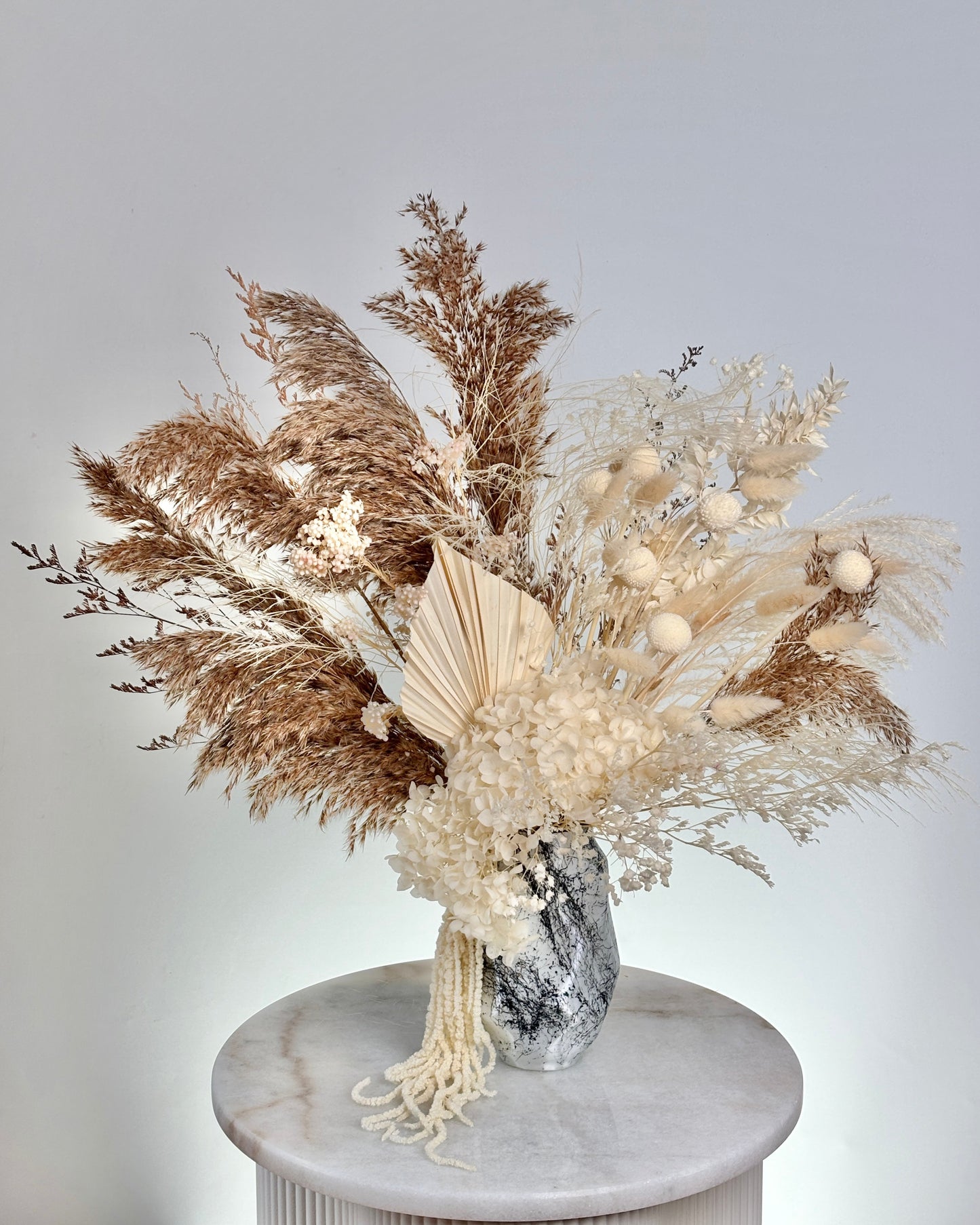 Decorative arrangement of dried pampas grass and flowers in a vase on a marble pedestal against a light grey background.
