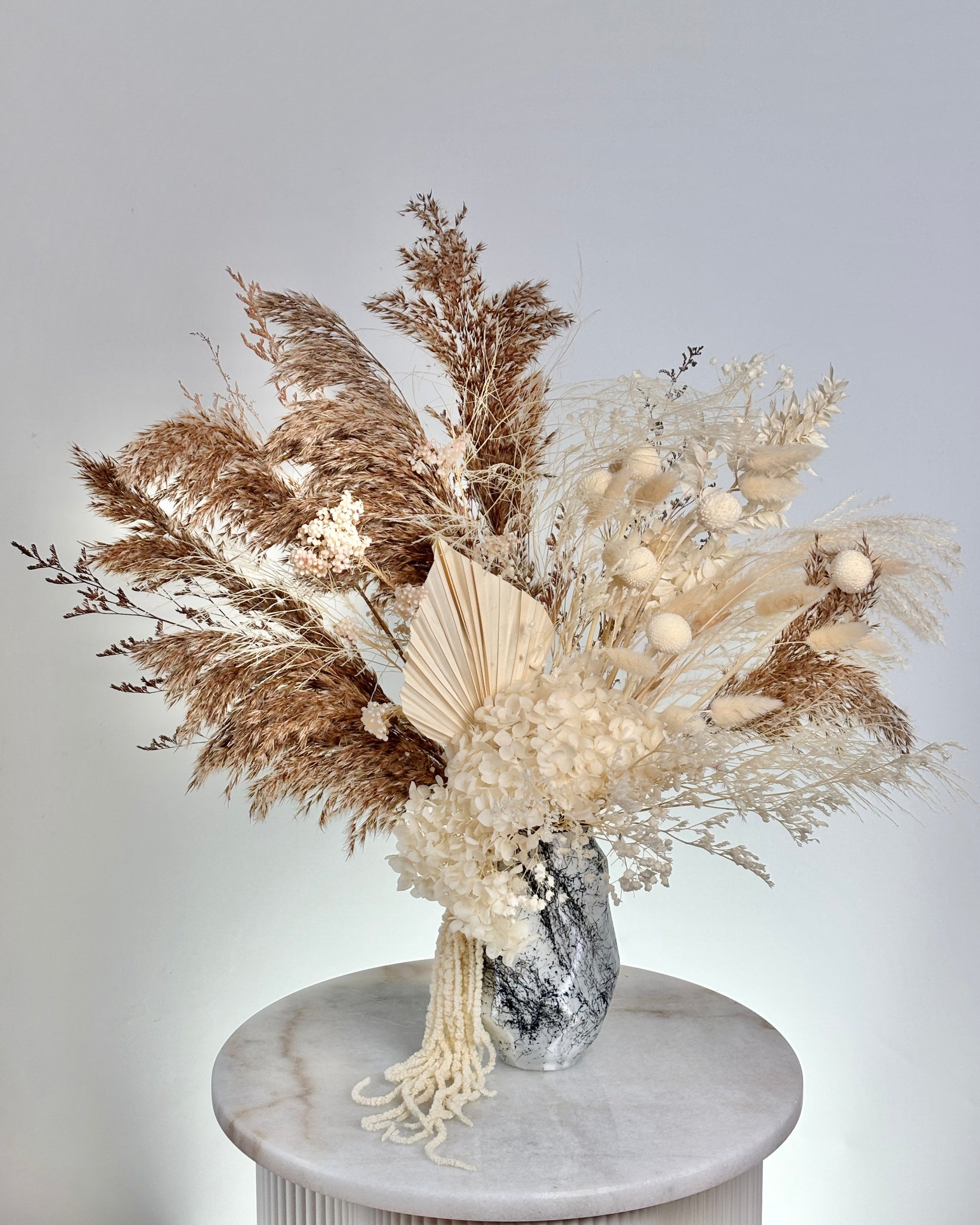 Decorative arrangement of dried pampas grass and flowers in a vase on a marble pedestal against a light grey background.