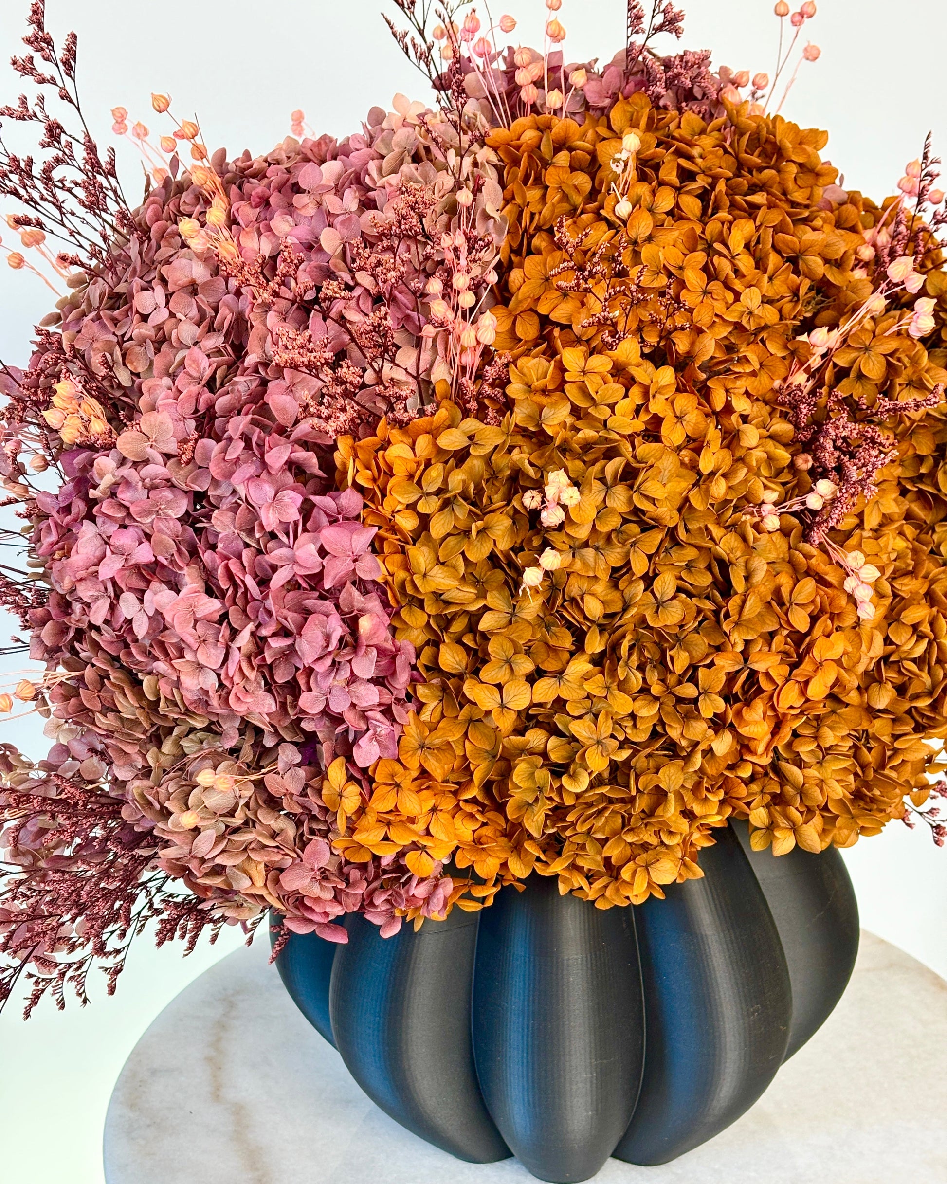 A dried floral arrangement featuring textured pink and golden-orange hydrangea blooms with small pale berries, presented in a dark matte ribbed vase on a white marble surface against a white background.