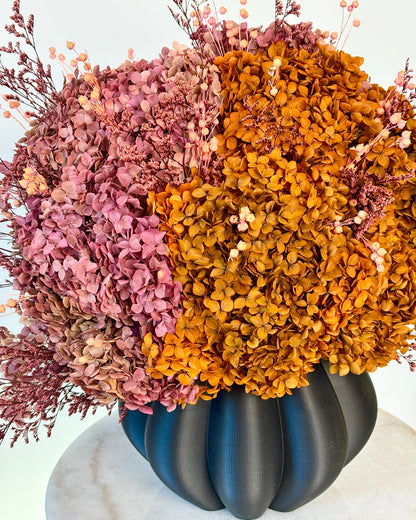 A dried floral arrangement featuring textured pink and golden-orange hydrangea blooms with small pale berries, presented in a dark matte ribbed vase on a white marble surface against a white background.
