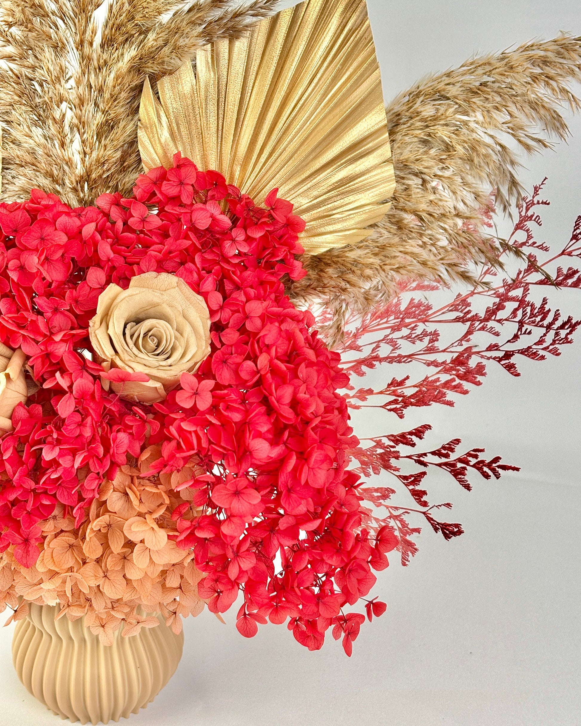 Decorative preserved floral arrangement with red flowers, a gold leaf, and pampas grass on a light background