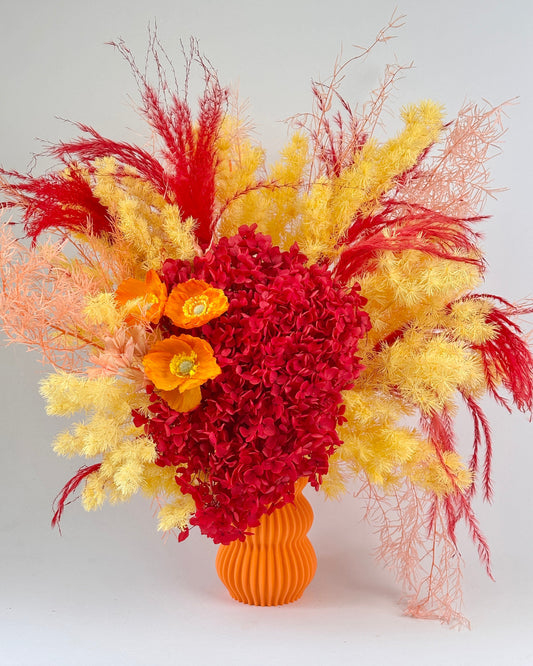 Colorful floral arrangement with red, yellow, and orange dried flowers in an orange vase on a light gray background