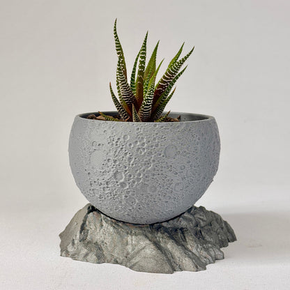 Cylindrical concrete planter with a small plant on a textured stone base against a light gray background