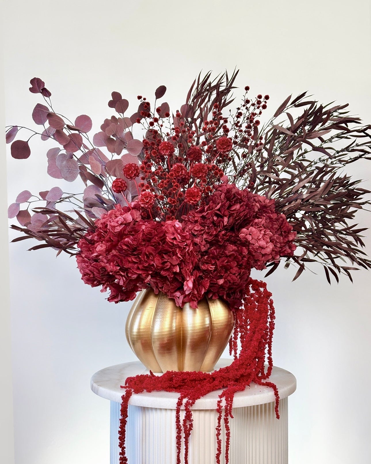 A dried floral arrangement in a ribbed gold vase, featuring deep red hydrangeas, burgundy berries, and dark burgundy foliage, displayed on a white pedestal against a light gray background.