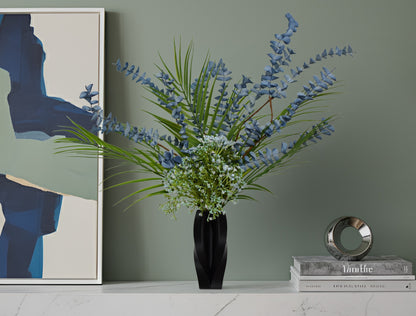Decorative arrangement of greenery and blue flowers in a black vase on a shelf with a book and decorative object against a green wall.