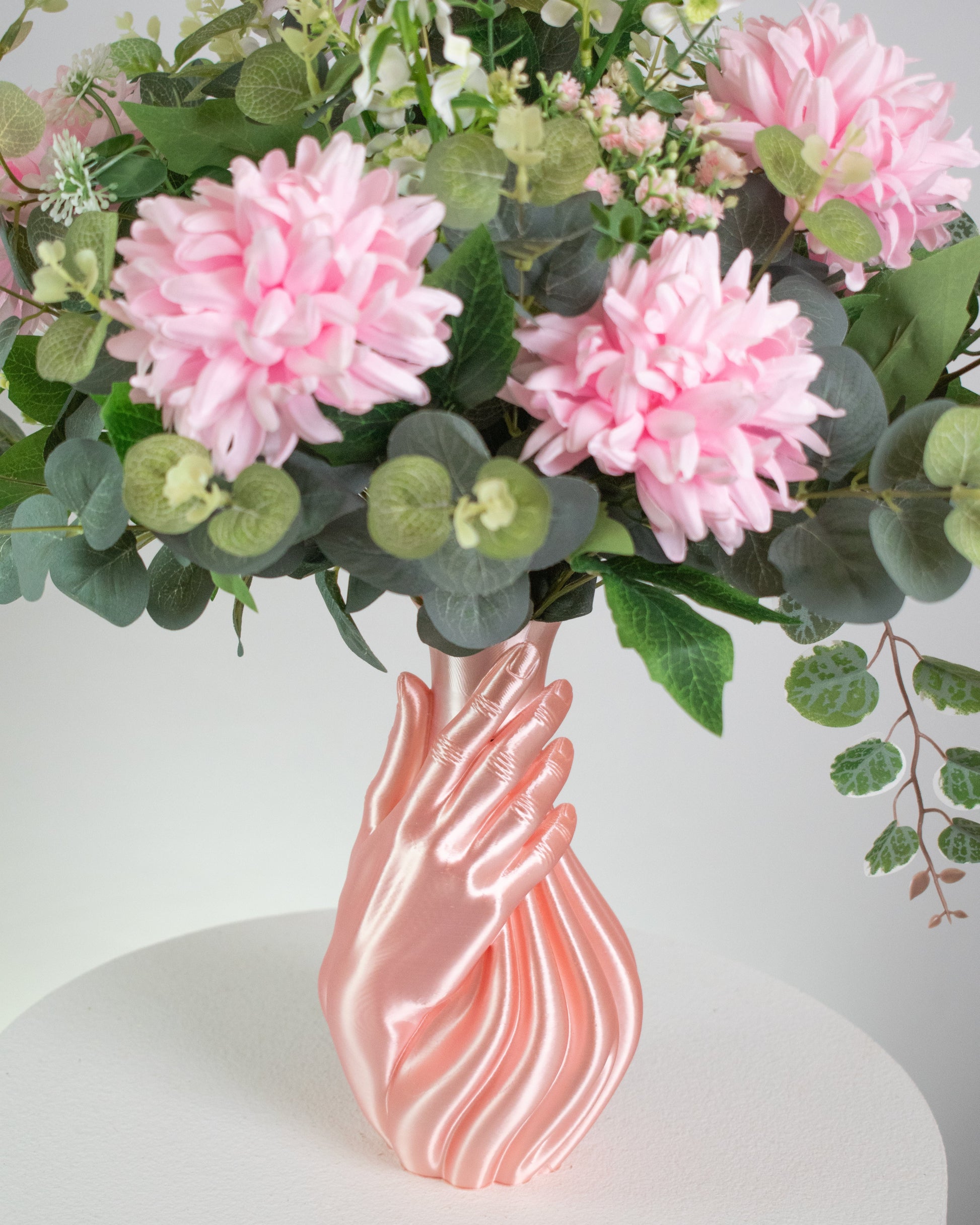 Artificial floral arrangement with pink and white blooms and lush green foliage, elegantly displayed in a sculptural pink hand-shaped vase on a neutral background.