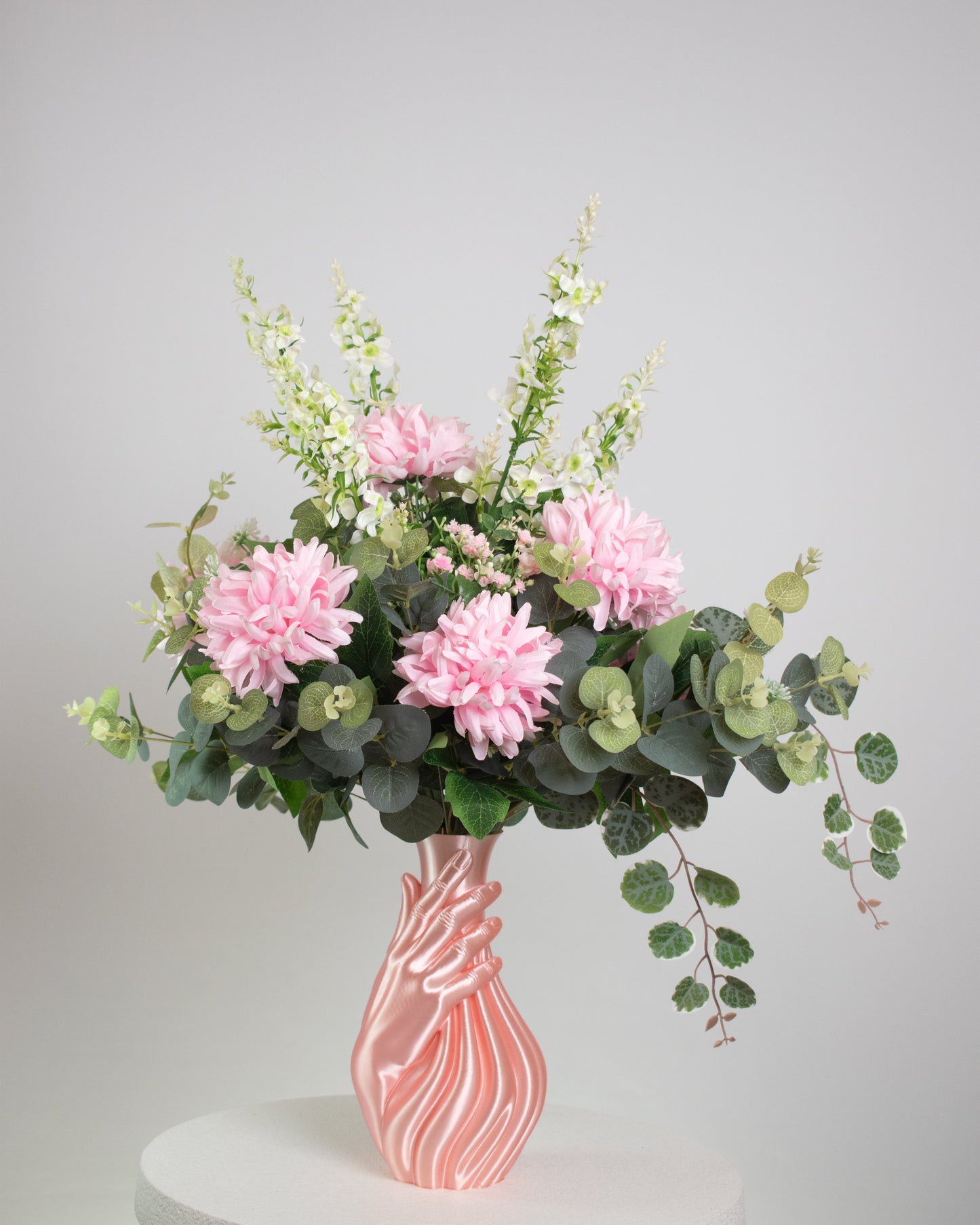 Artificial floral arrangement with pink and white blooms and lush green foliage, elegantly displayed in a sculptural pink hand-shaped vase on a neutral background.