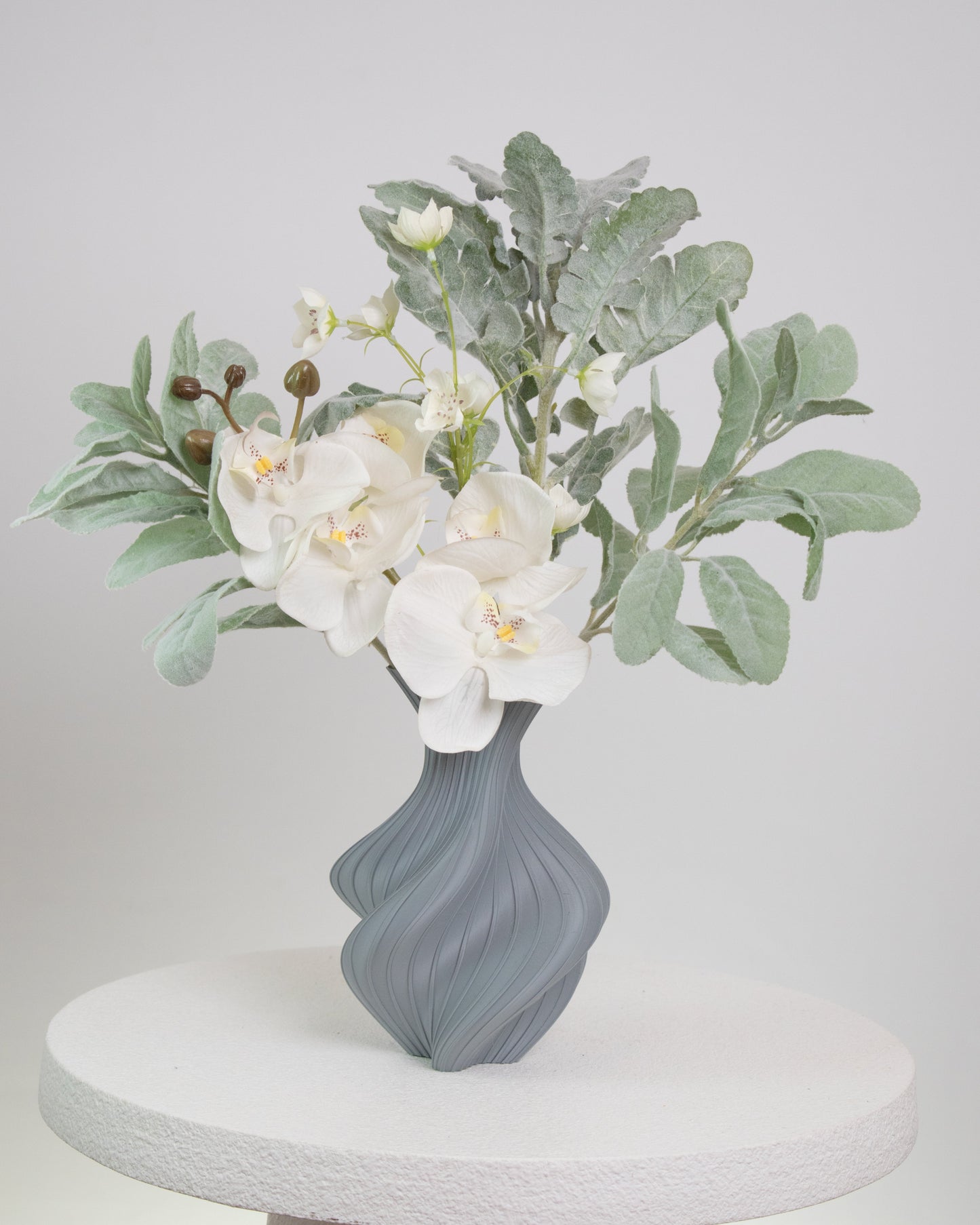 Artificial floral arrangement featuring white orchids and silver-green foliage in a sculptural grey vase with curved lines, displayed on a round white pedestal.
